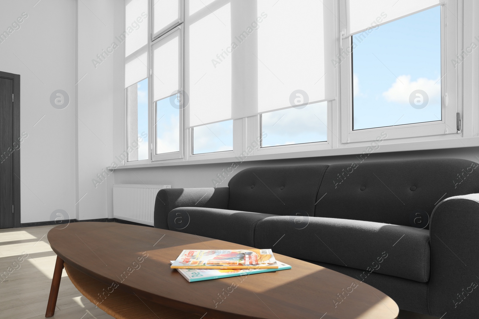 Photo of Comfortable sofa and table near large window with white roller blinds indoors