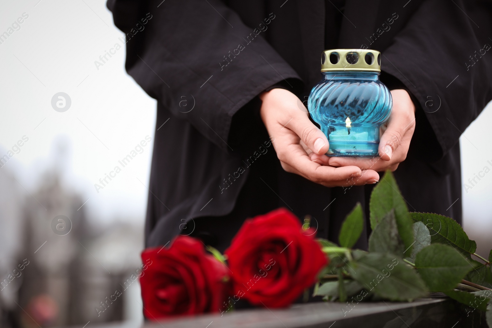 Photo of Woman holding candle near tombstone with red roses outdoors, closeup. Funeral ceremony