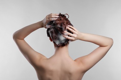 Young woman washing her hair with shampoo on light grey background, back view