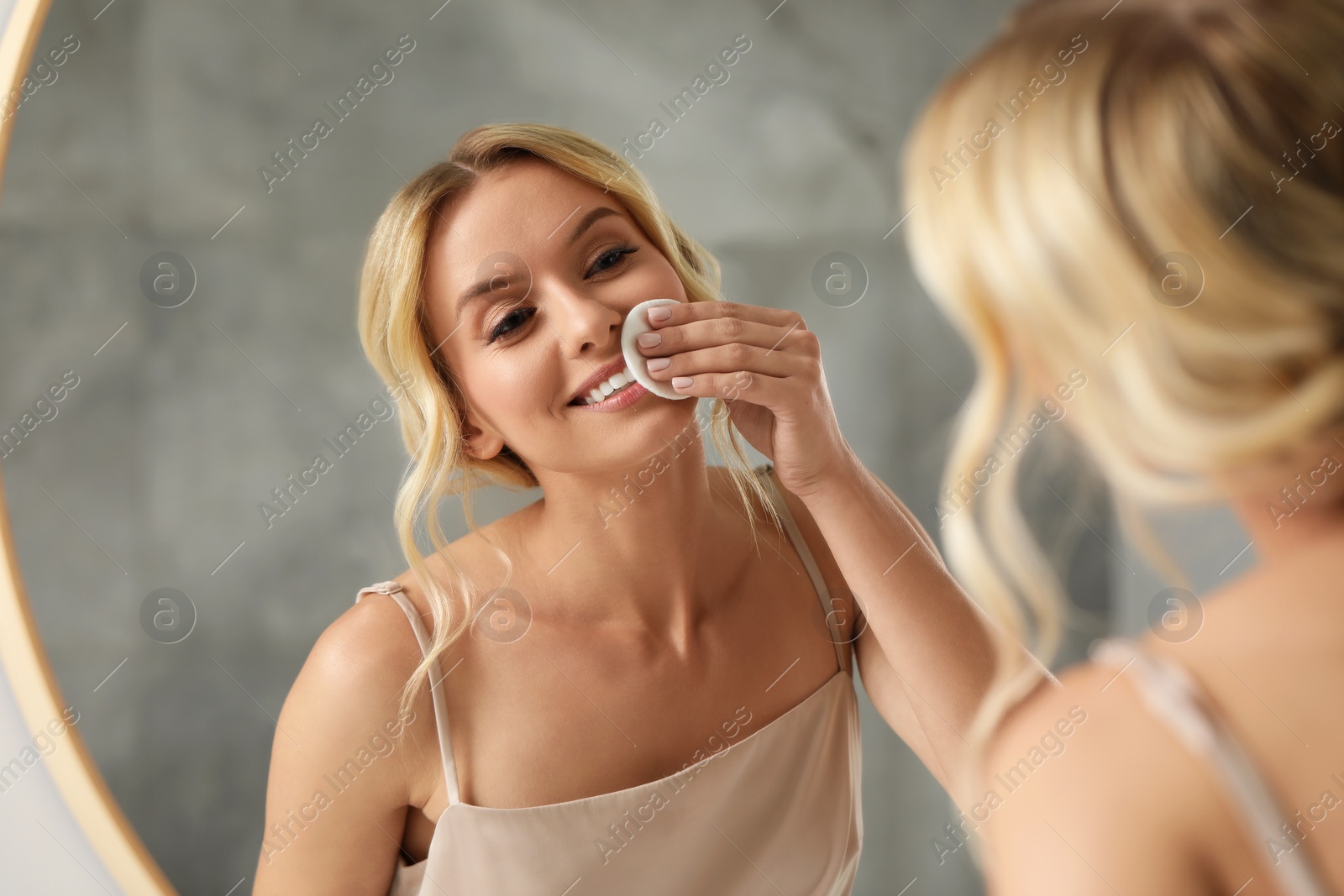 Photo of Smiling woman removing makeup with cotton pad in front of mirror in bathroom