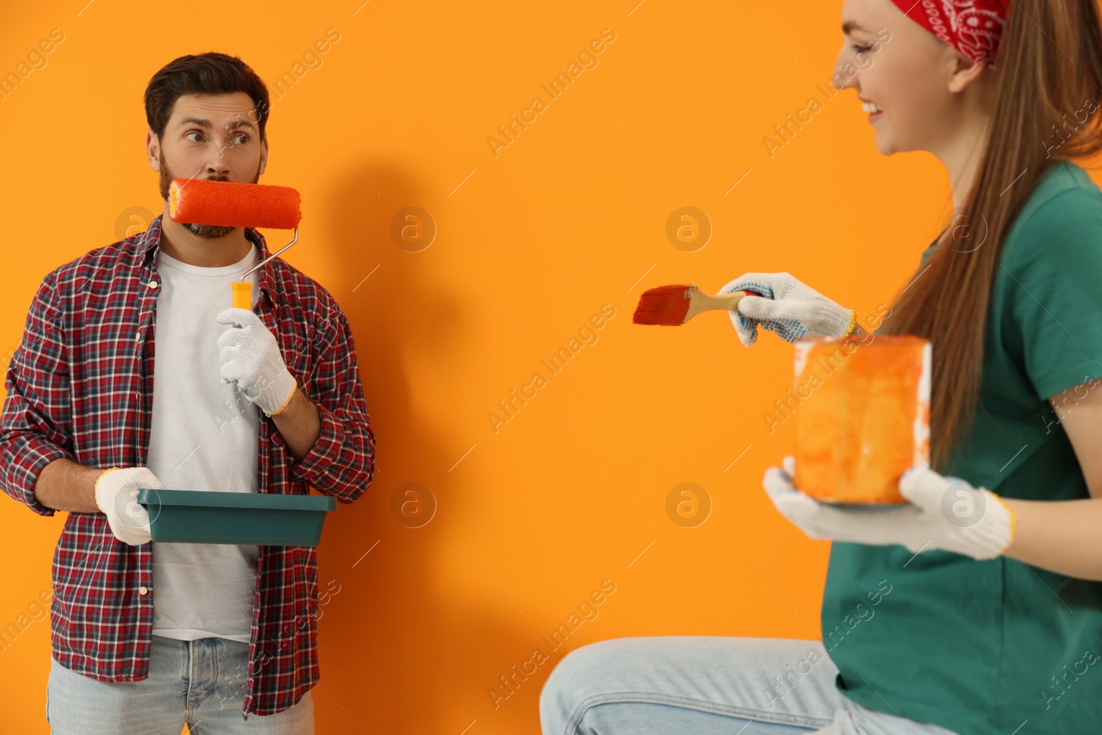 Photo of Man making funny face expression with roller and woman holding can of dye near freshly painted orange wall. Interior design