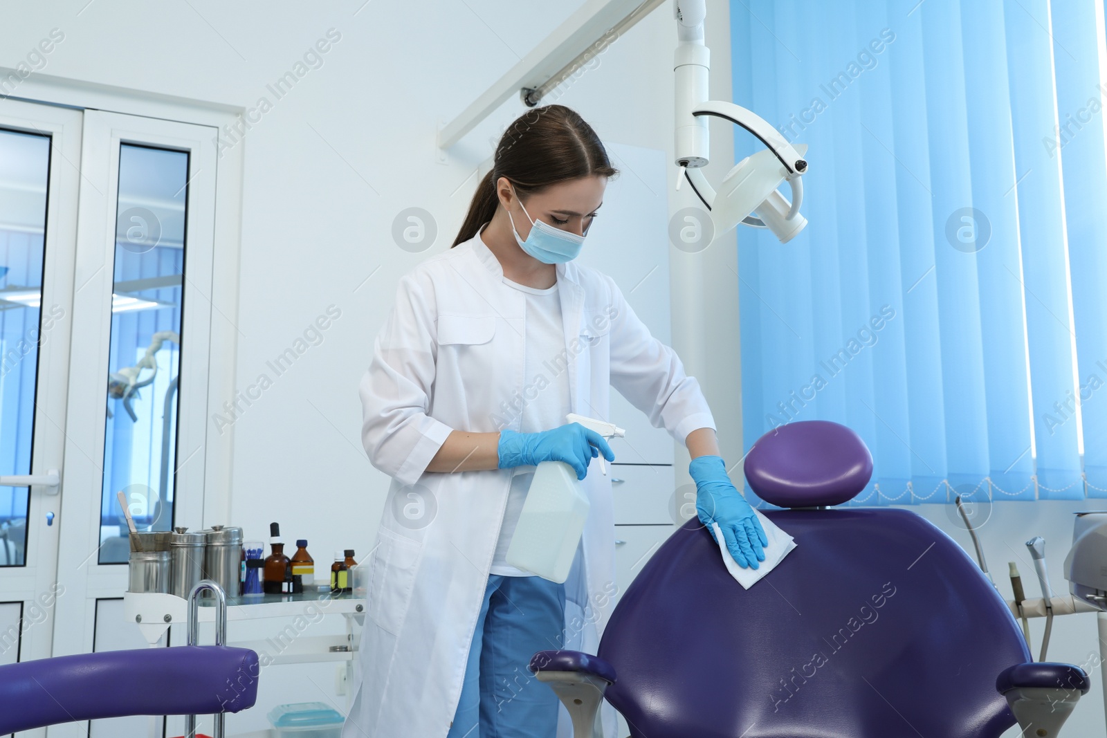 Photo of Professional dentist in white coat and medical mask cleaning workplace with antiseptic indoors