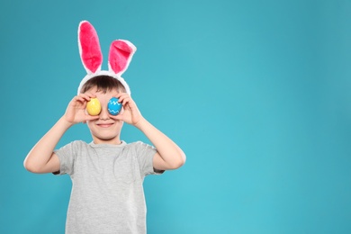 Little boy in bunny ears headband holding Easter eggs near eyes on color background, space for text