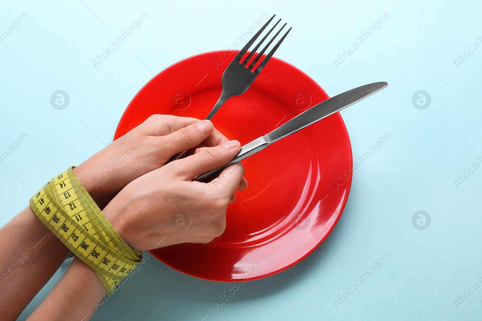 Photo of Diet concept. Woman holding cutlery in hands tied with measuring tape over empty plate on light blue background, above view