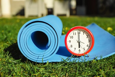 Alarm clock and fitness mat on green grass outdoors. Morning exercise