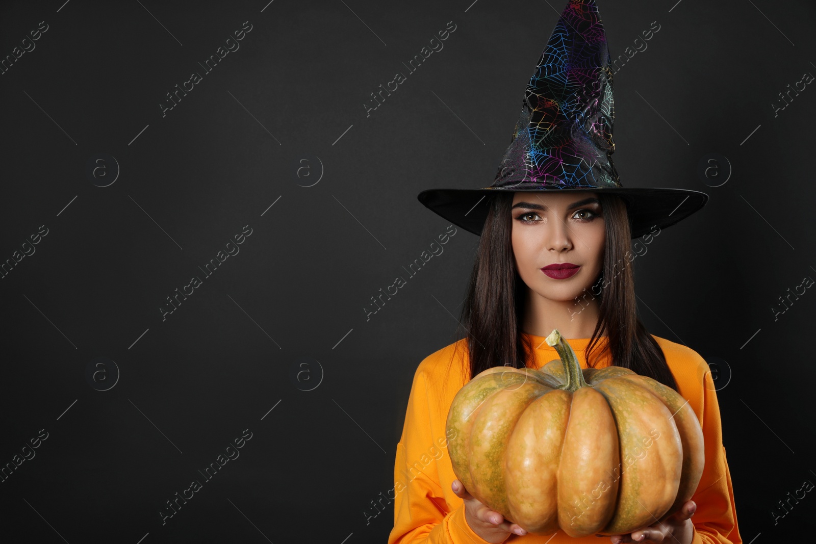 Photo of Beautiful woman wearing witch costume with pumpkin for Halloween party on black background, space for text