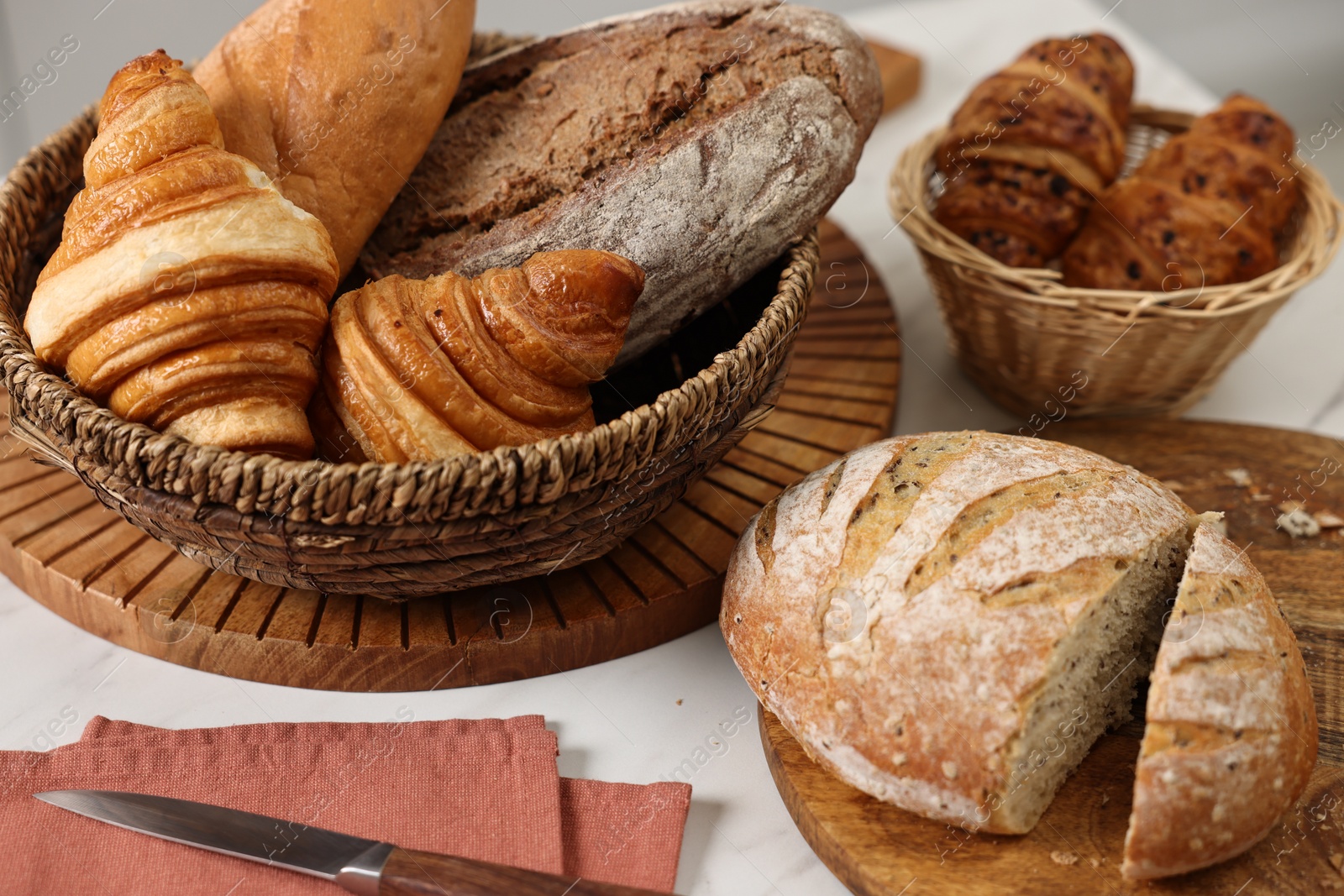 Photo of Wicker bread basket with freshly baked loaves and knife on white marble table in kitchen, closeup