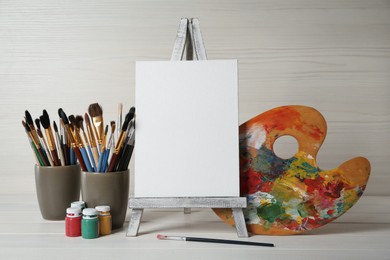 Photo of Easel with blank canvas, brushes, paints and palette on white wooden table