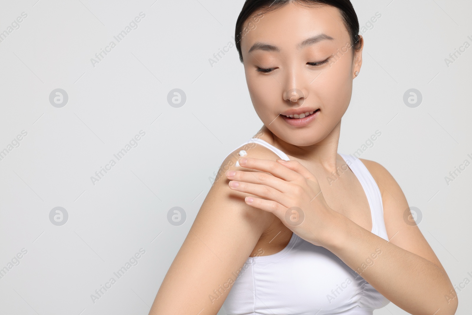 Photo of Beautiful young Asian woman applying body cream onto shoulder on light grey background, space for text