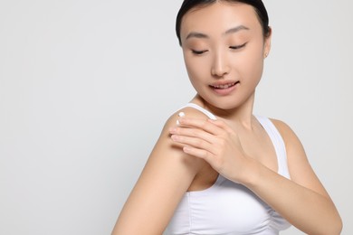 Photo of Beautiful young Asian woman applying body cream onto shoulder on light grey background, space for text