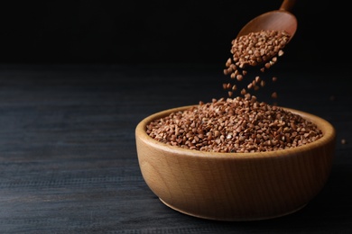 Photo of Pouring buckwheat grains from spoon into wooden bowl on grey table. Space for text