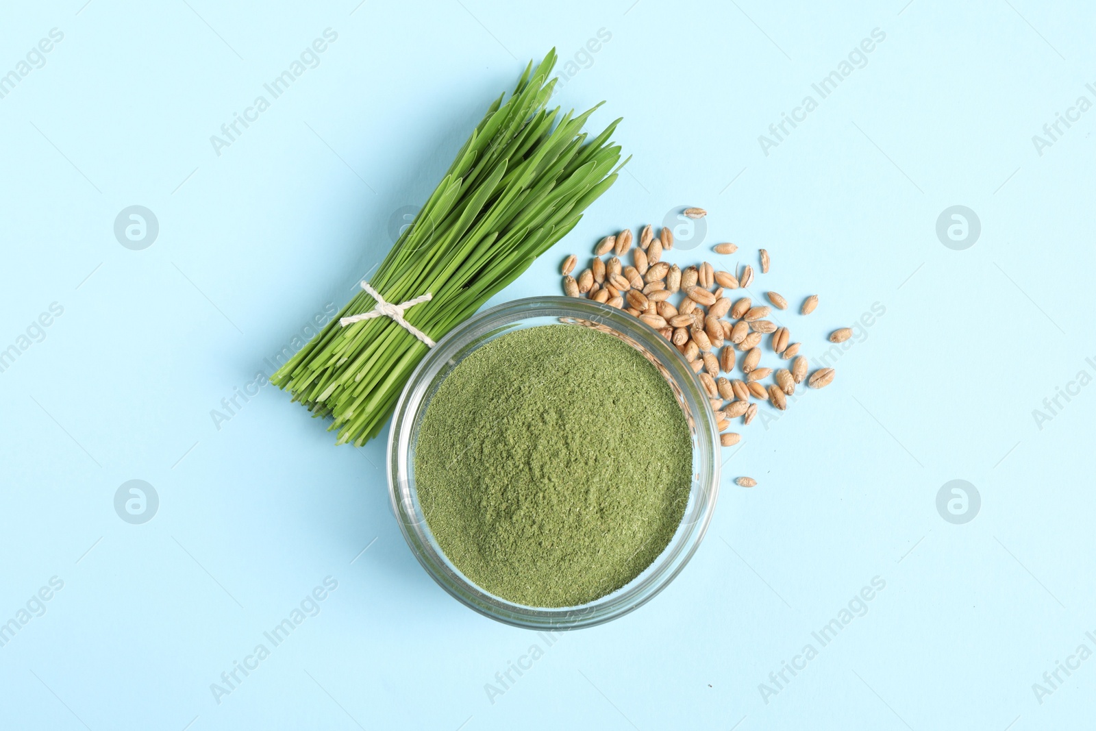 Photo of Wheat grass powder in glass bowl, seeds and fresh sprouts on light blue table, flat lay