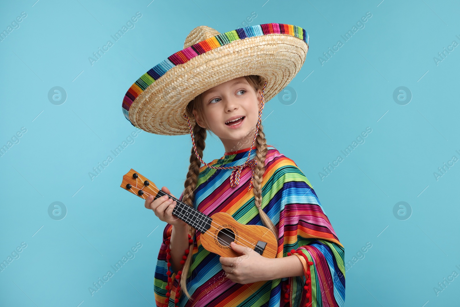 Photo of Cute girl in Mexican sombrero hat and poncho playing ukulele on light blue background