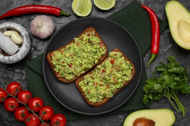 Slices of bread with tasty guacamole and ingredients on black textured table, flat lay