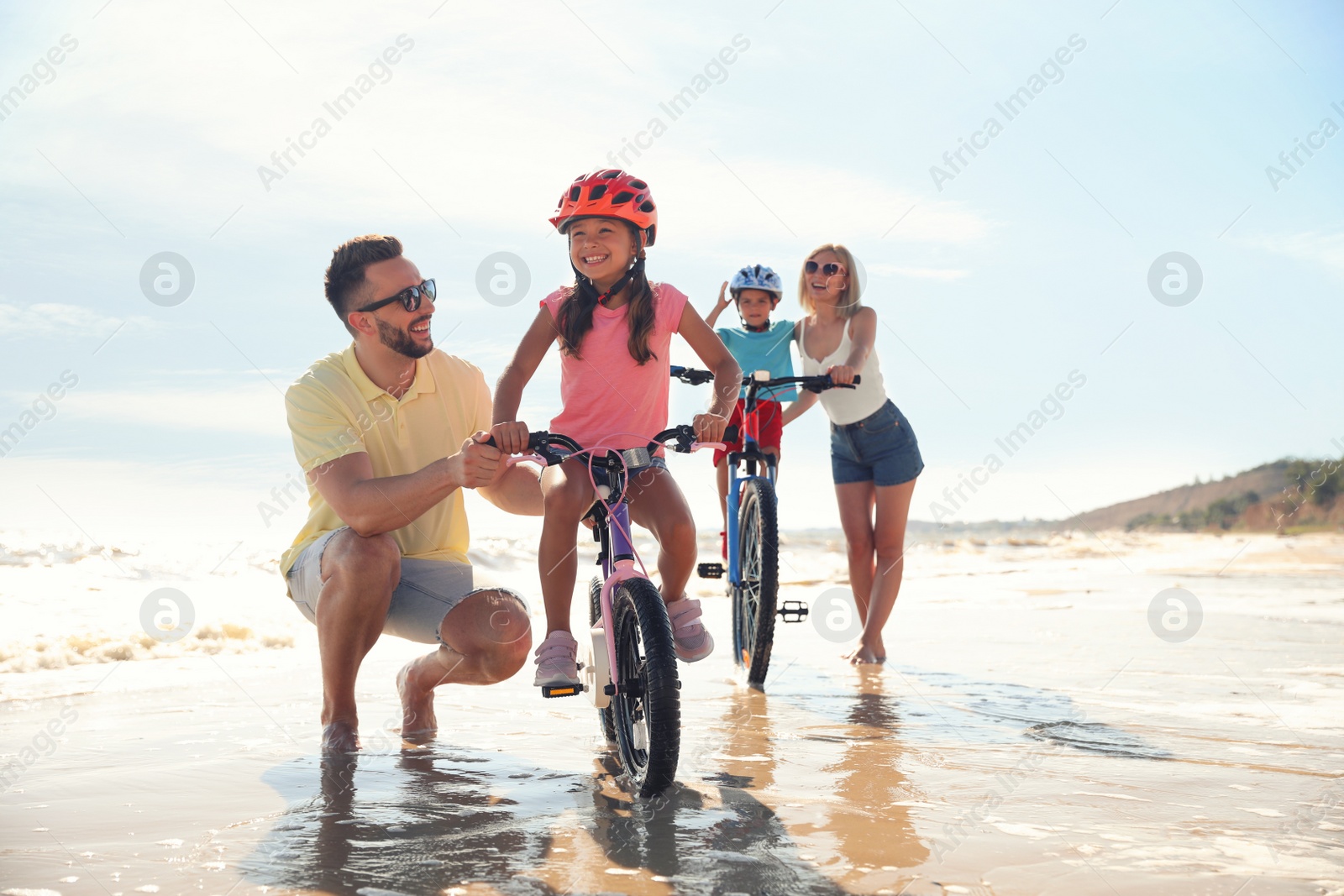 Photo of Happy parents teaching children to ride bicycles on sandy beach near sea