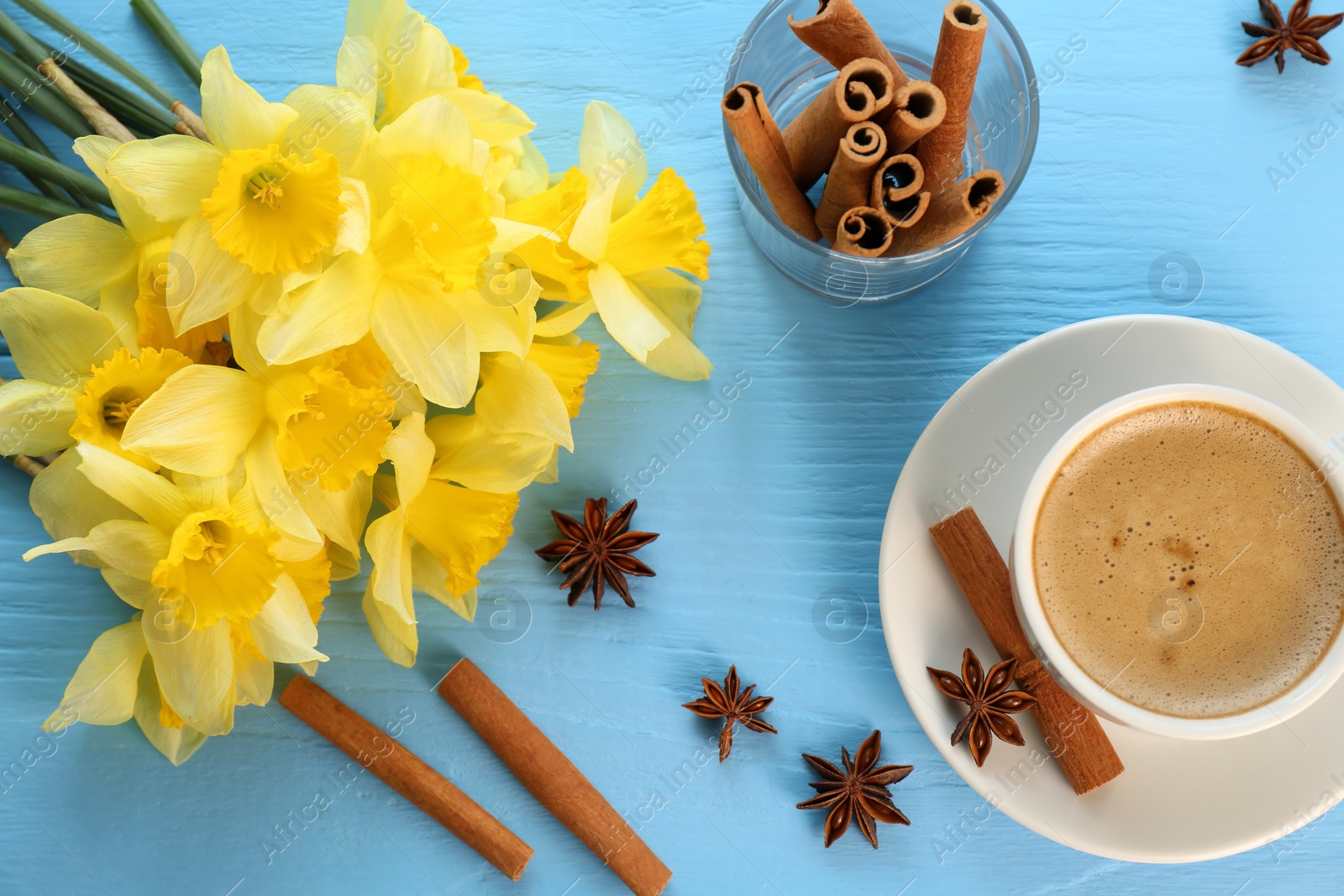 Photo of Beautiful yellow daffodils, cup of coffee, cinnamon sticks and anise stars on light blue wooden table, flat lay