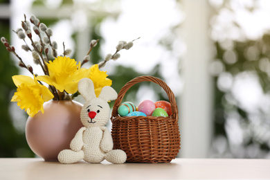 Photo of Festive composition with Easter eggs in wicker basket and bunny on table against blurred window, space for text