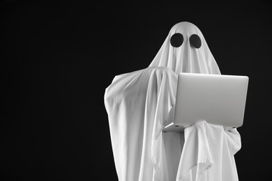 Photo of Creepy ghost. Person in white sheet with laptop on black background, space for text