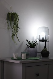 Photo of Stylish lamp, candles and green plants on grey chest of drawers indoors. Interior design