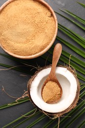 Photo of Spoon with coconut sugar, fruit, bowl and palm leaves on dark textured table, flat lay