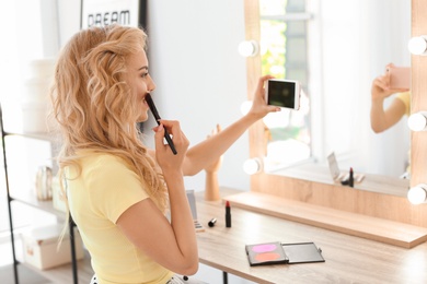 Photo of Beauty blogger filming makeup tutorial with smartphone in front of mirror at dressing table indoors