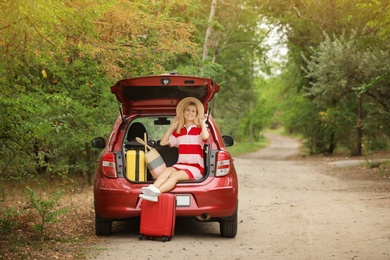 Photo of Happy woman sitting in car trunk with suitcases near forest. Space for text