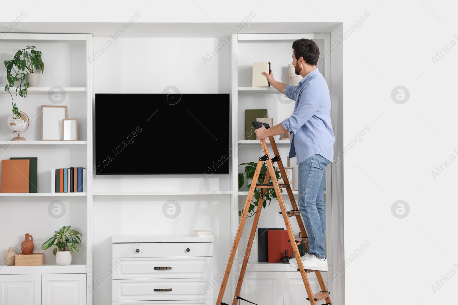 Photo of Man on wooden folding ladder taking book from shelf at home