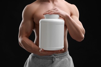 Young man with muscular body holding jar of protein powder on black background, closeup