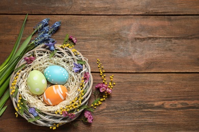 Flat lay composition of wicker nest with painted Easter eggs and flowers on wooden table, space for text