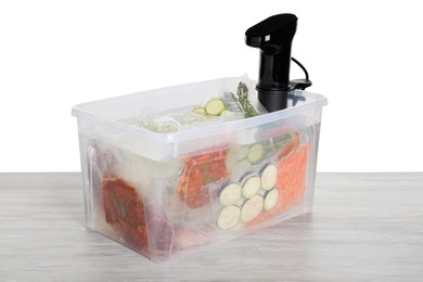 Photo of Sous vide cooker and vacuum packed food products in box on wooden table against white background. Thermal immersion circulator