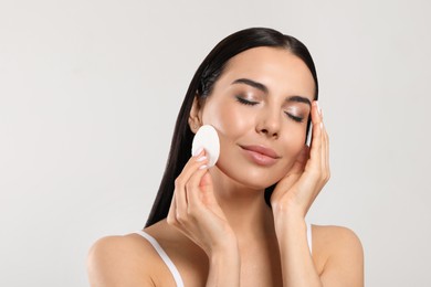 Young woman using cotton pad with micellar water on light grey background