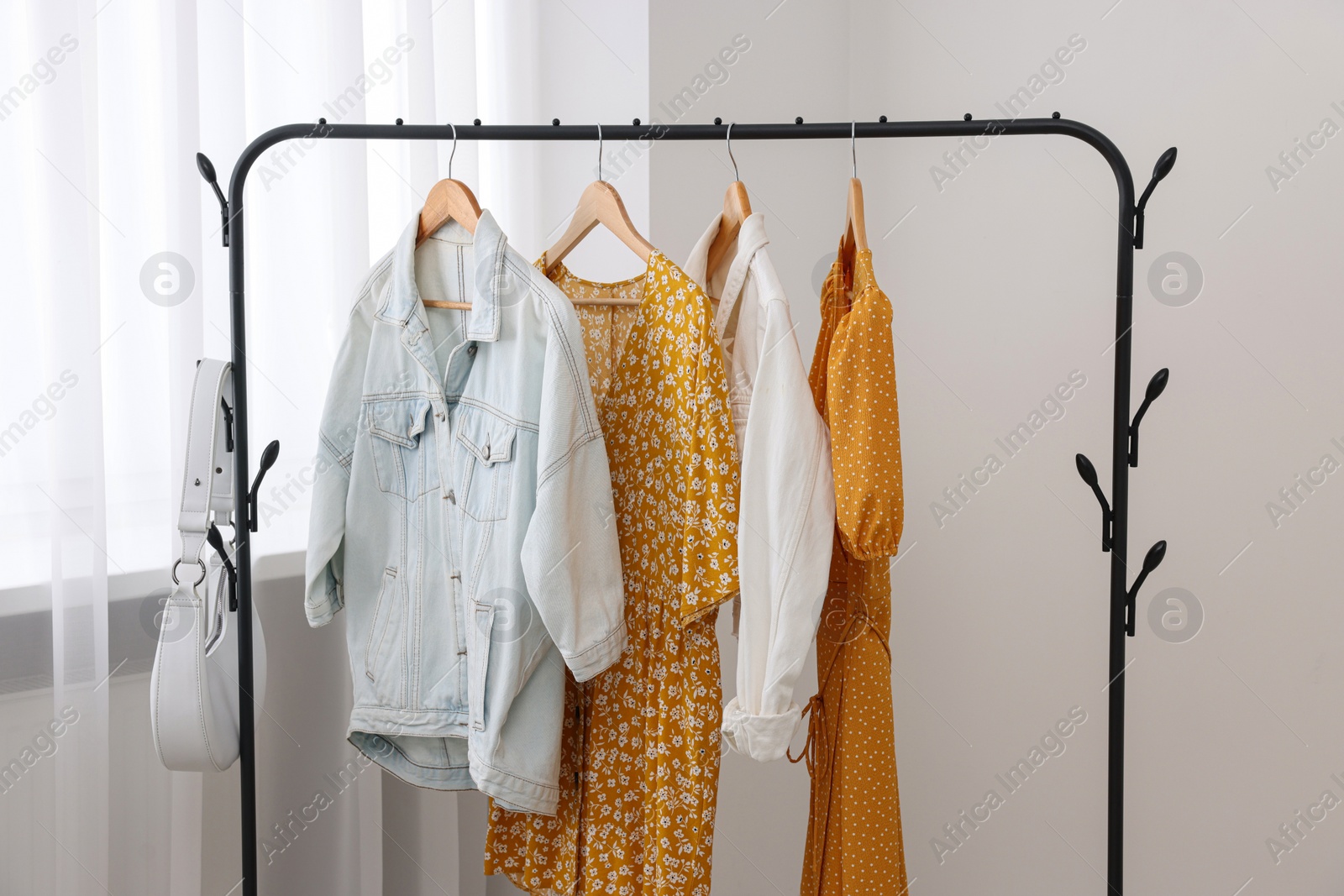 Photo of Rack with bag and stylish women's clothes on hangers near light wall indoors