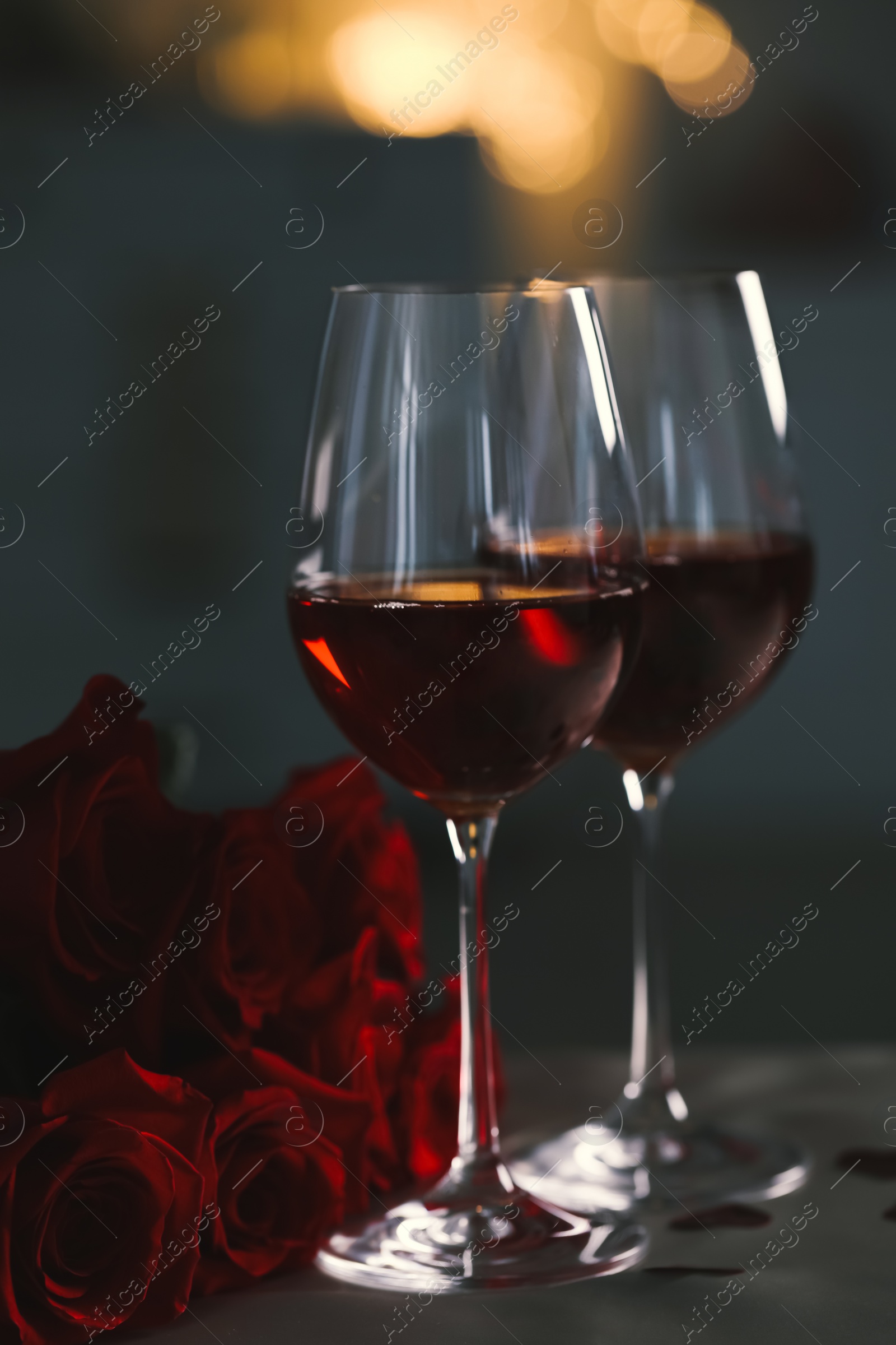 Photo of Glasses of red wine and rose flowers on grey table against blurred lights. Romantic atmosphere