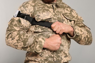 Photo of Soldier in military uniform applying medical tourniquet on arm against light grey background, closeup
