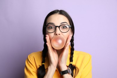 Fashionable young woman with braids blowing bubblegum on lilac background