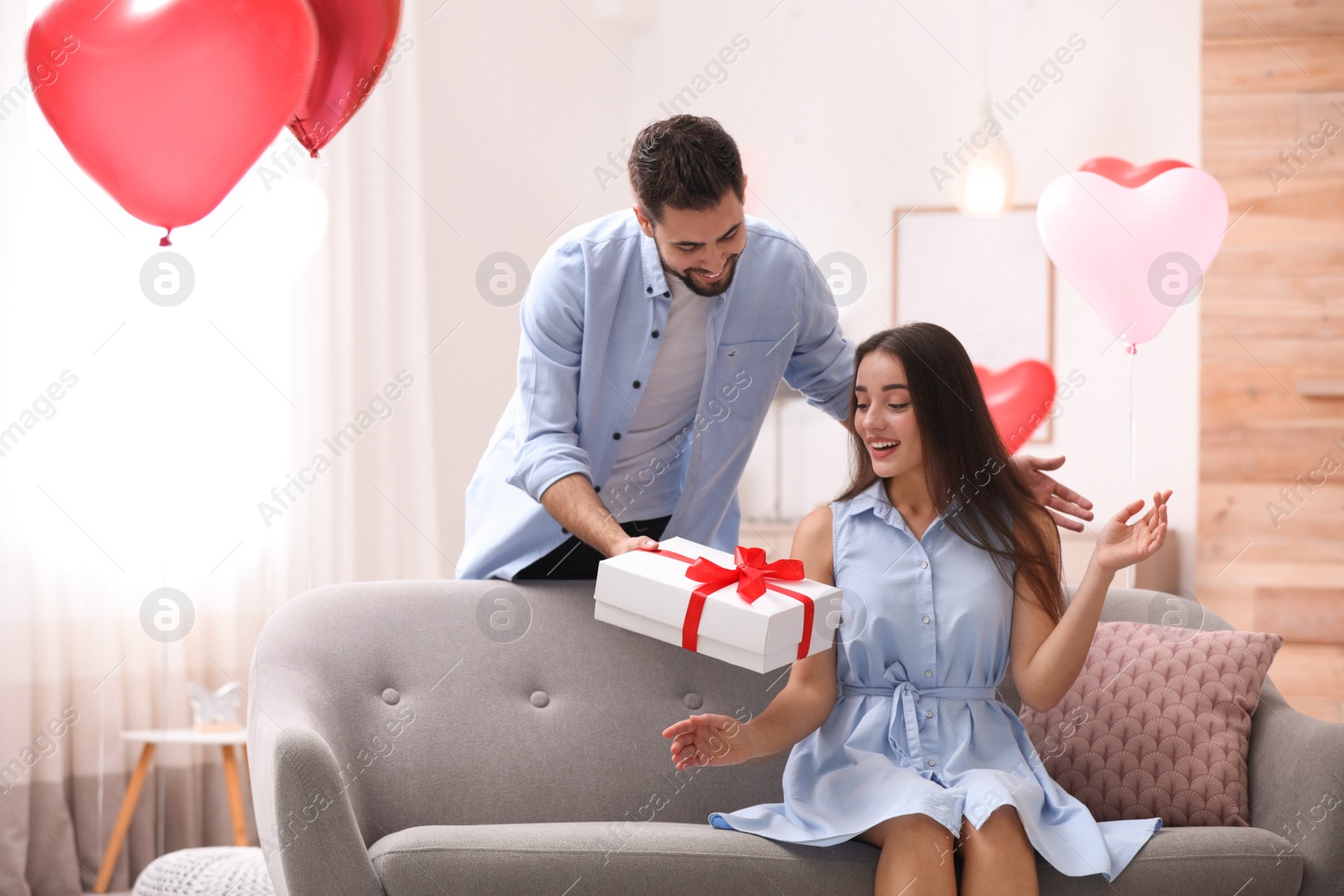 Photo of Young man presenting gift to his girlfriend in living room decorated with heart shaped balloons. Valentine's day celebration