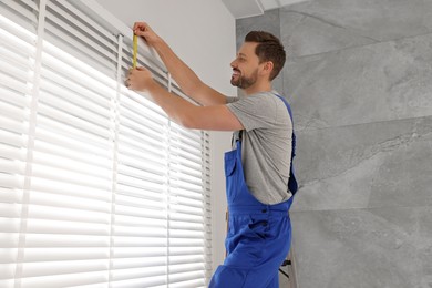 Worker in uniform using measuring tape while installing horizontal window blinds indoors