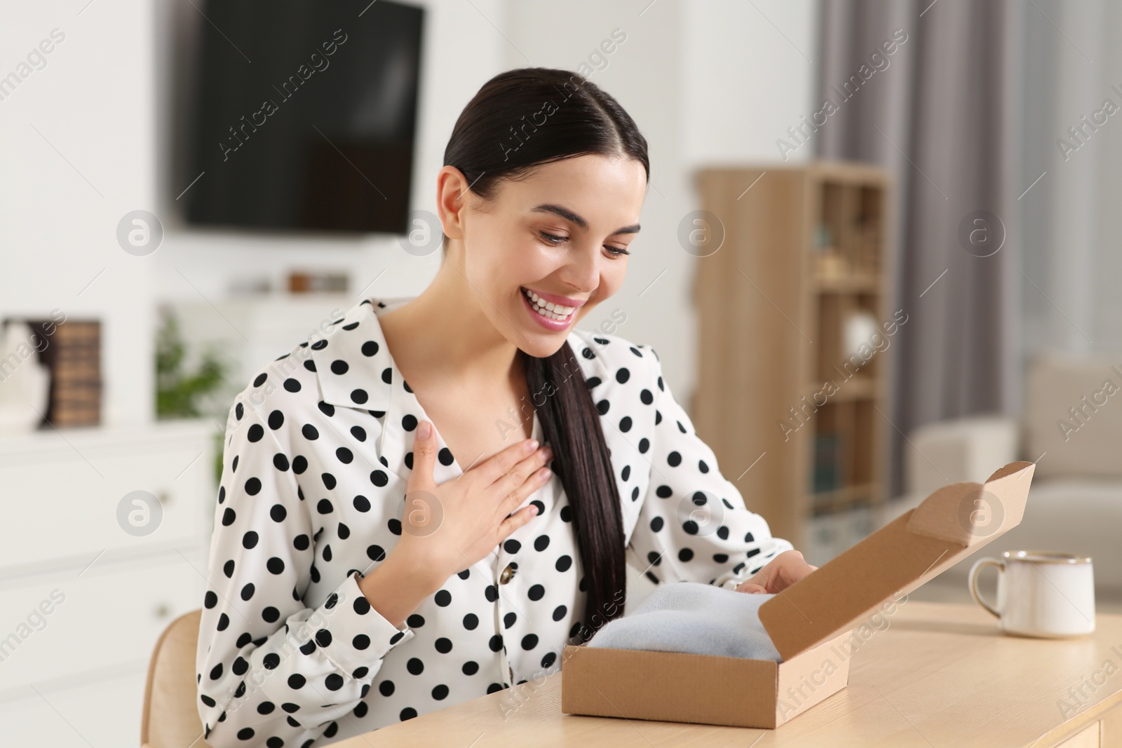 Photo of Happy young woman opening parcel at table indoors. Internet shopping