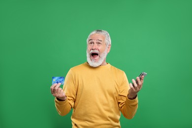 Photo of Shocked senior man with credit card and smartphone on green background. Be careful - fraud