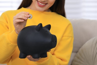 Young woman putting coin into piggy bank indoors, closeup