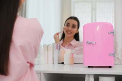 Photo of Woman doing face massage at dressing table with cosmetic refrigerator indoors