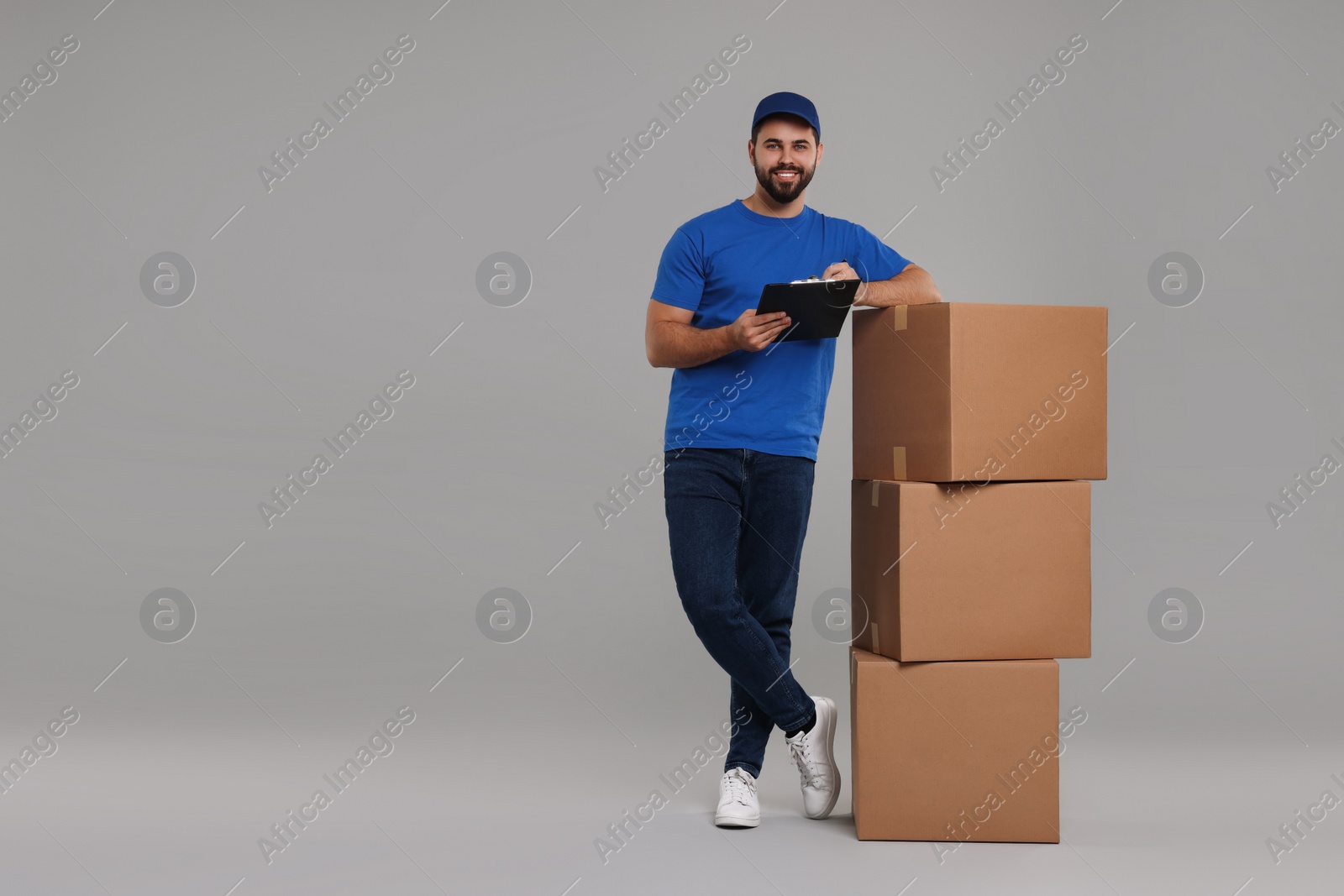 Photo of Happy young courier with stack of parcels and clipboard on grey background, space for text