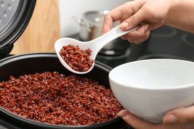 Photo of Woman putting brown rice into bowl from multi cooker in kitchen, closeup