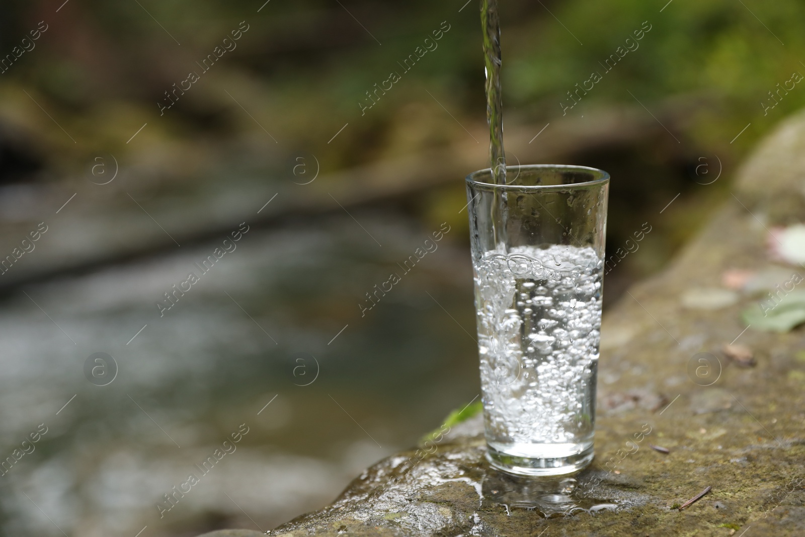 Photo of Fresh water pouring into glass on stone near stream. Space for text