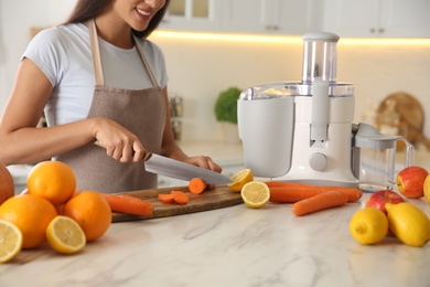 Young woman cutting fresh carrot for juice at table in kitchen, closeup