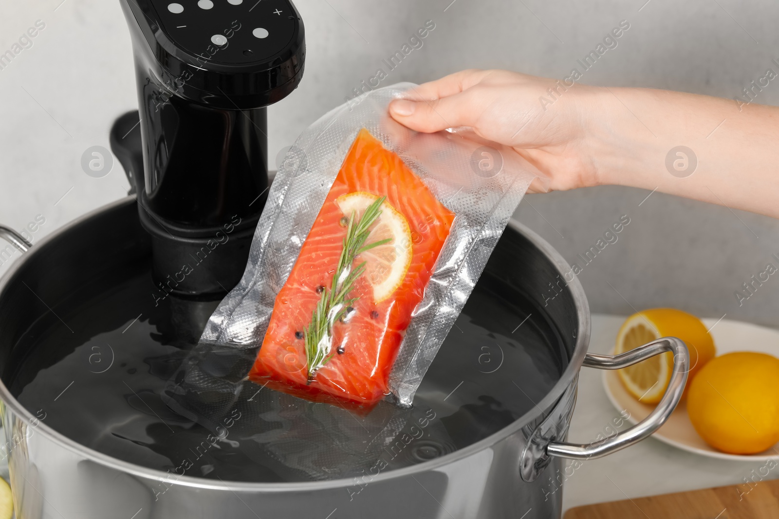 Photo of Woman putting vacuum packed salmon into pot with sous vide cooker in kitchen, closeup. Thermal immersion circulator