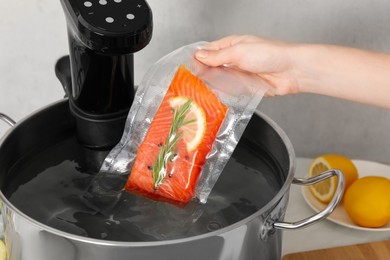 Photo of Woman putting vacuum packed salmon into pot with sous vide cooker in kitchen, closeup. Thermal immersion circulator