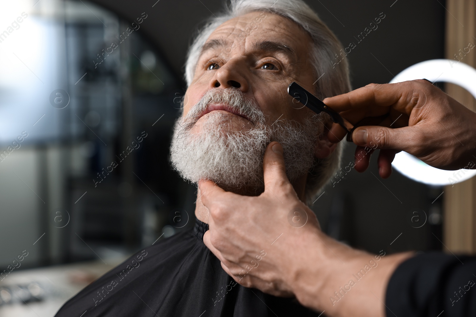 Photo of Professional barber shaving client's beard with blade in barbershop