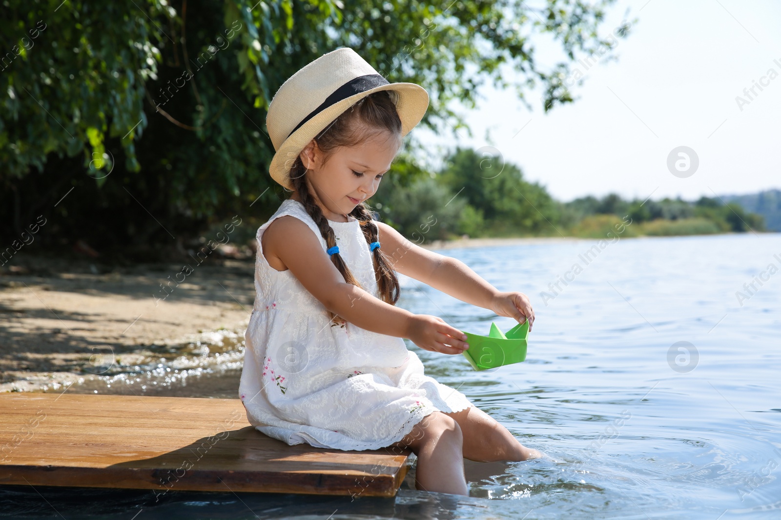 Photo of Cute little girl playing with paper boat on wooden pier near river