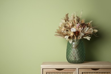 Photo of Beautiful dried flower bouquet in glass vase on wooden table near olive wall. Space for text